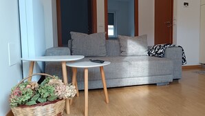 A living room with a sofa, two small tables, a basket of flowers, and a wooden floor. - Olive Tree House- Vacation Rentals (Riva del Garda)