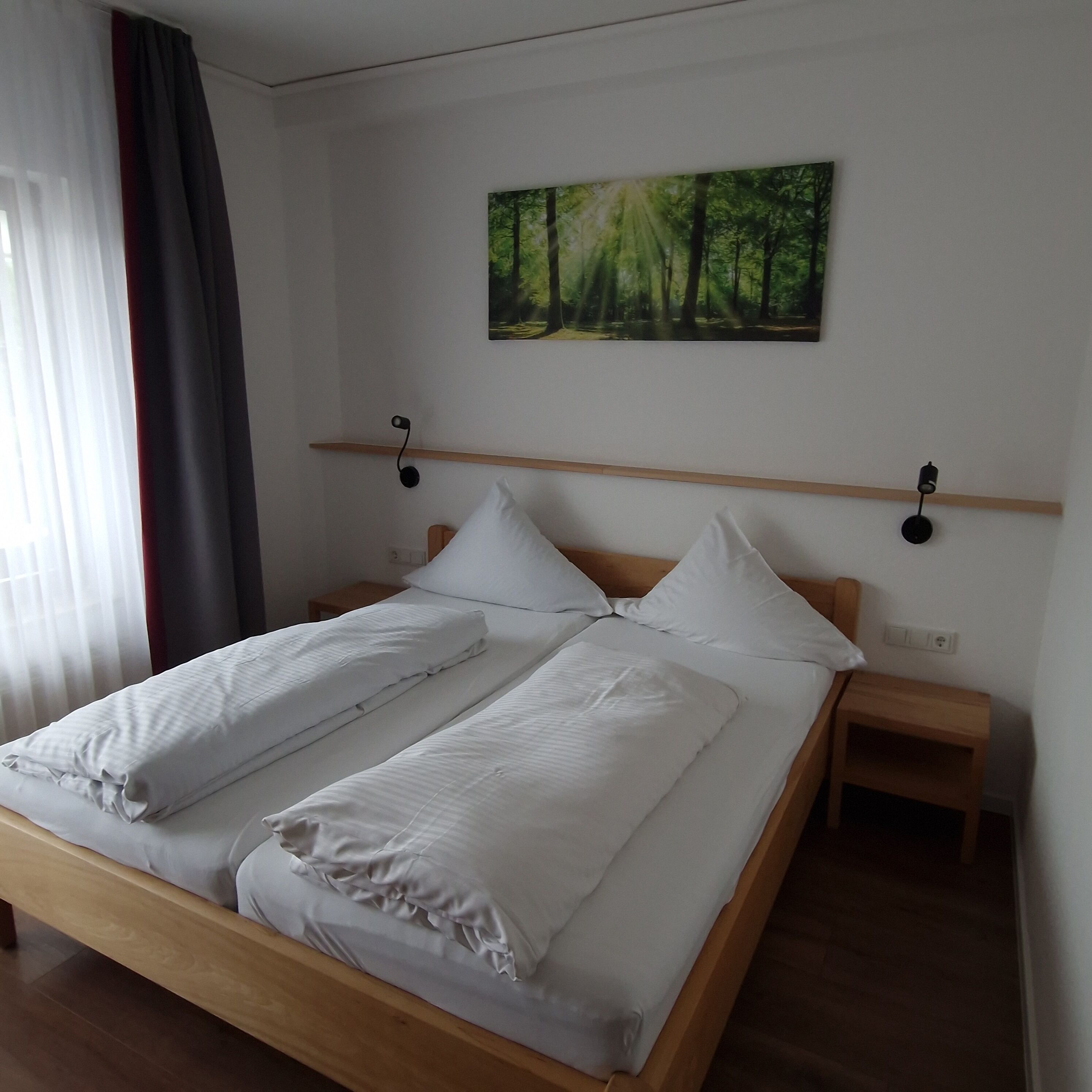 A neatly made double bed with white linens, two bedside tables, a wall-mounted shelf, and a forest-themed painting above the bed.