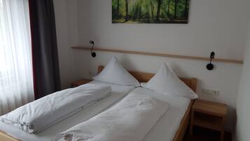 A neatly made double bed with white linens, two bedside tables, a wall-mounted shelf, and a forest-themed painting above the bed.