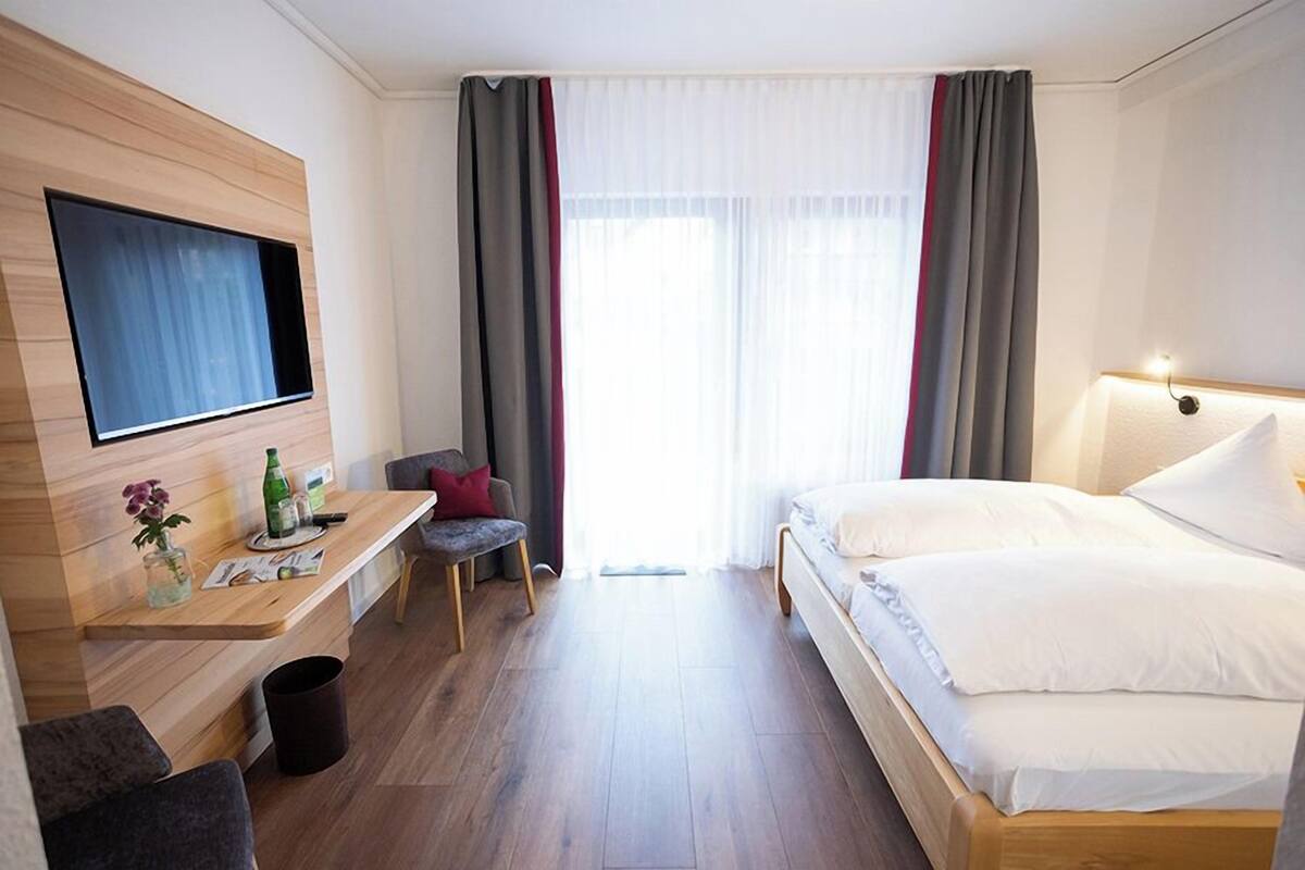 Una habitación de hotel moderna con un escritorio de madera, un televisor de pantalla plana, una cama con sábanas blancas y un ventanal con cortinas.