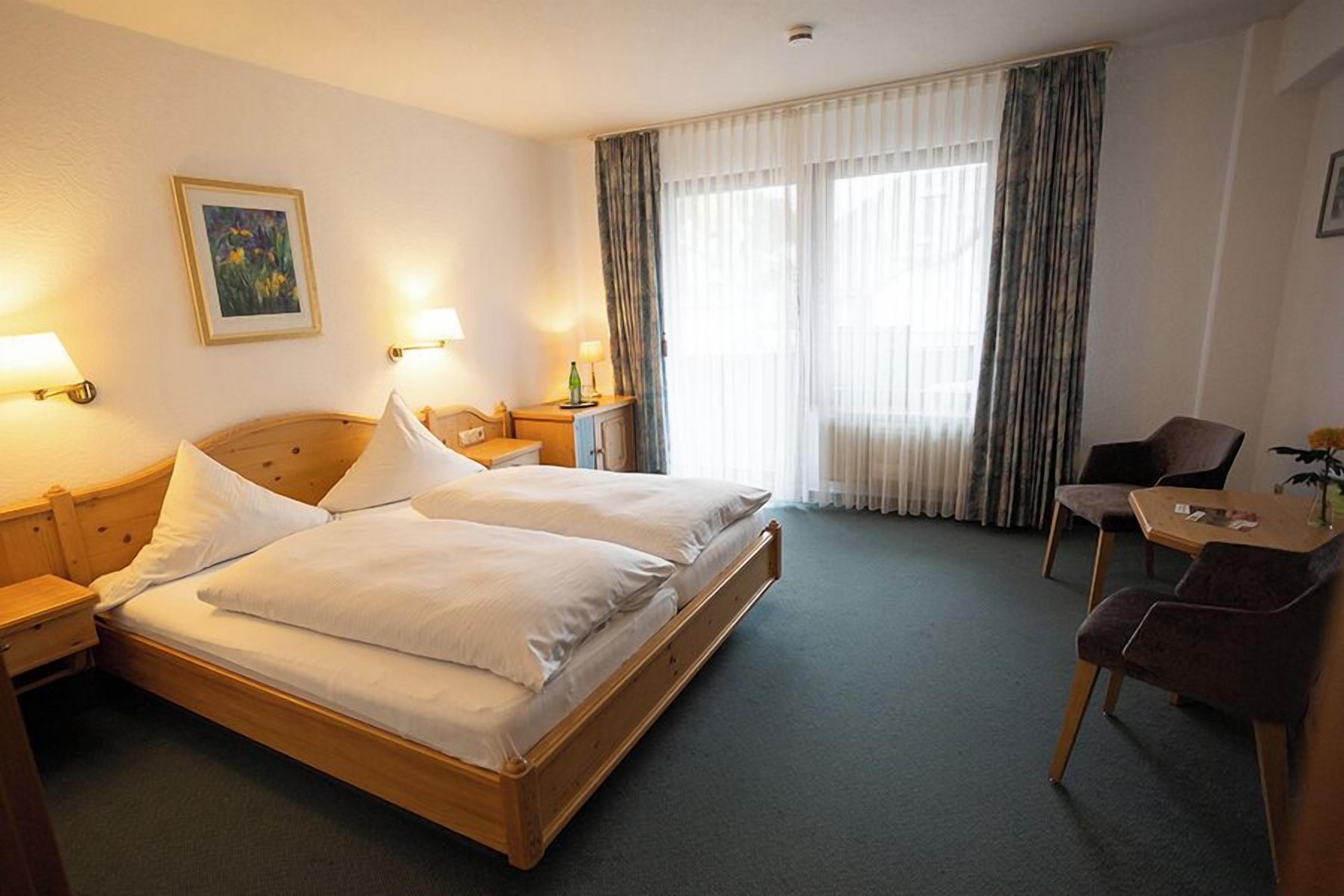 Quarto de hotel com uma cama de casal, duas mesas de cabeceira com luminárias, uma mesinha com uma cadeira e uma janela grande com cortinas.