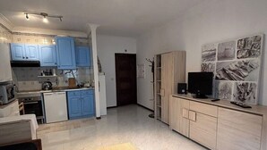 Una cocina compacta con armarios azules, un refrigerador pequeño y un microondas. Hay un mueble de madera con un televisor de pantalla plana encima.