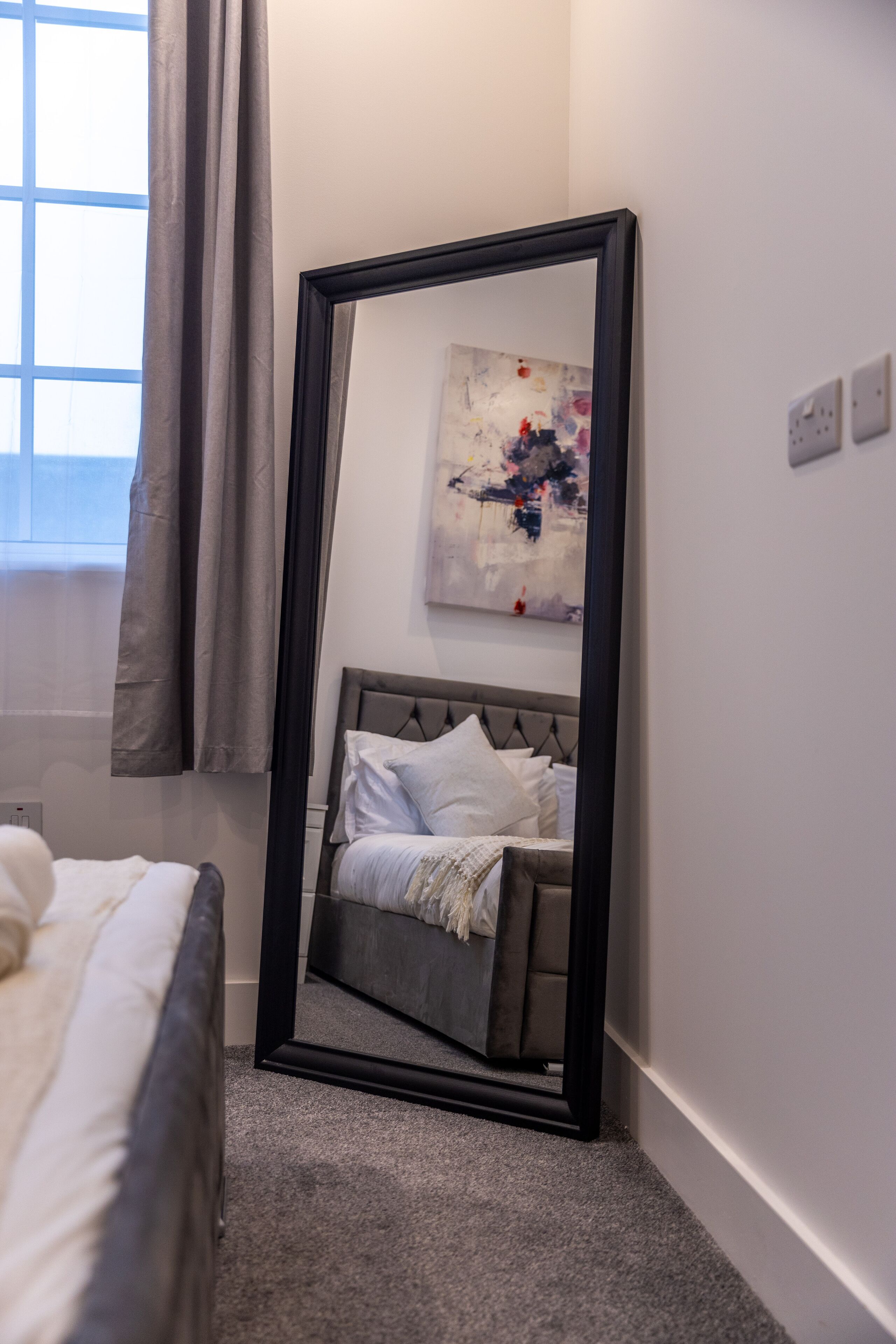 A bedroom with a large mirror, a bed, a headboard, and a painting on the wall.