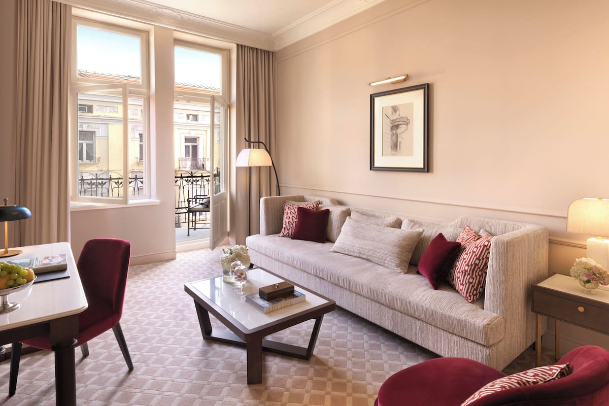 A modern hotel room with a sofa, armchairs, a coffee table, and a dining area with a table and chairs.
