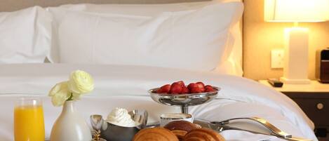 A neatly made bed with white linens, a bedside table with a lamp, and a breakfast tray with croissants, strawberries, and a glass of orange juice.