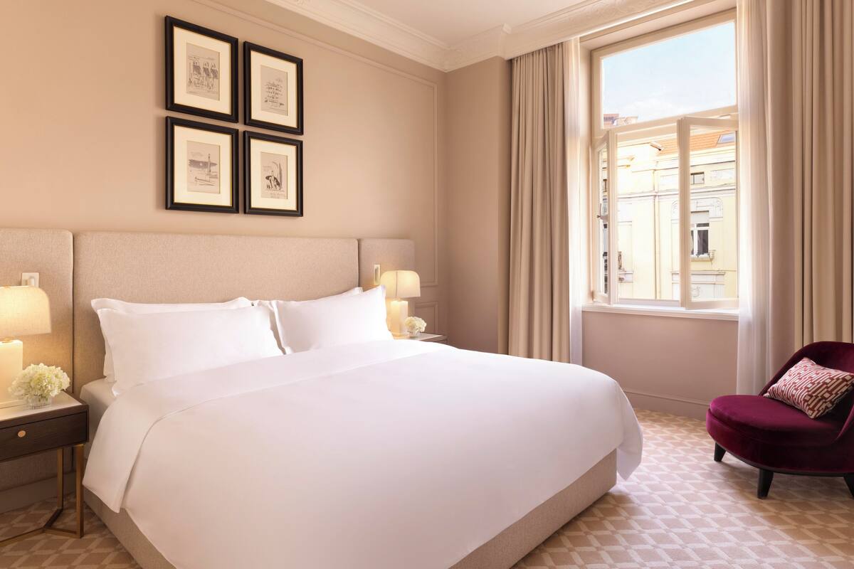 A hotel room with a large bed, two bedside lamps, a red armchair, and a window with curtains.
