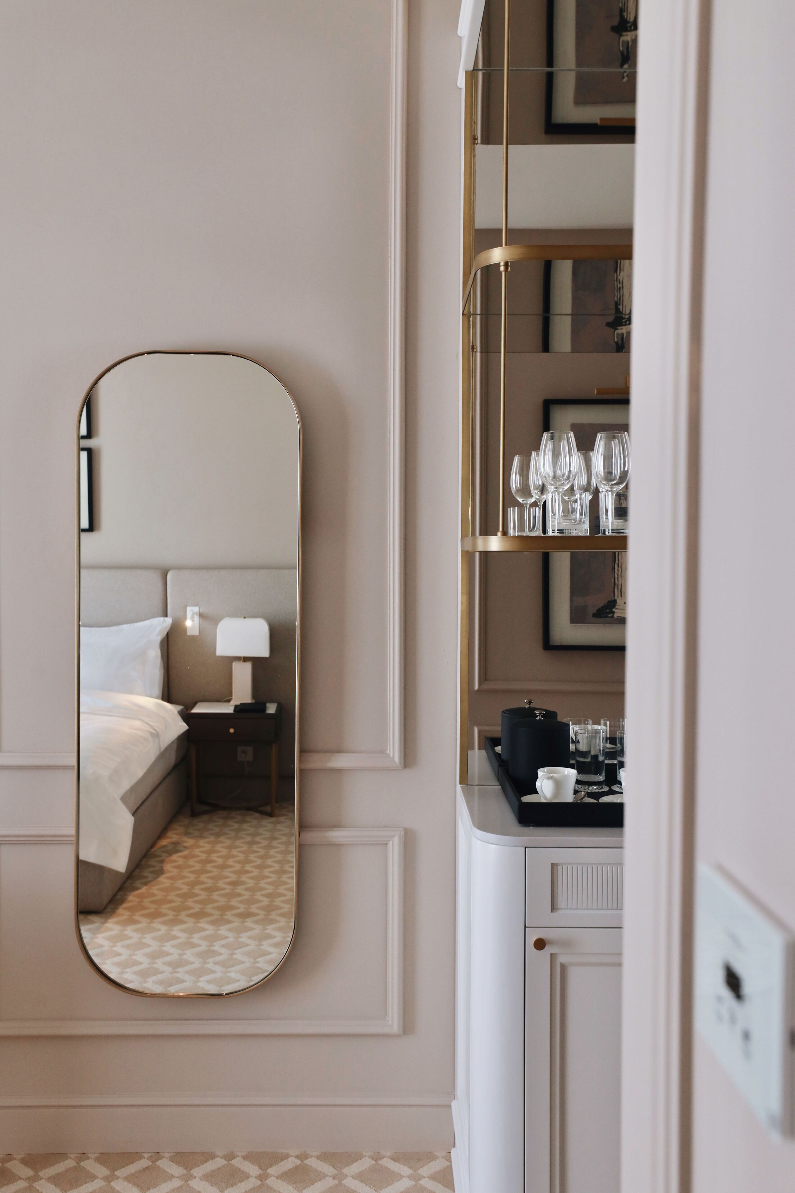 A hotel room with a large mirror, a bed, a nightstand with a lamp, and a minibar.