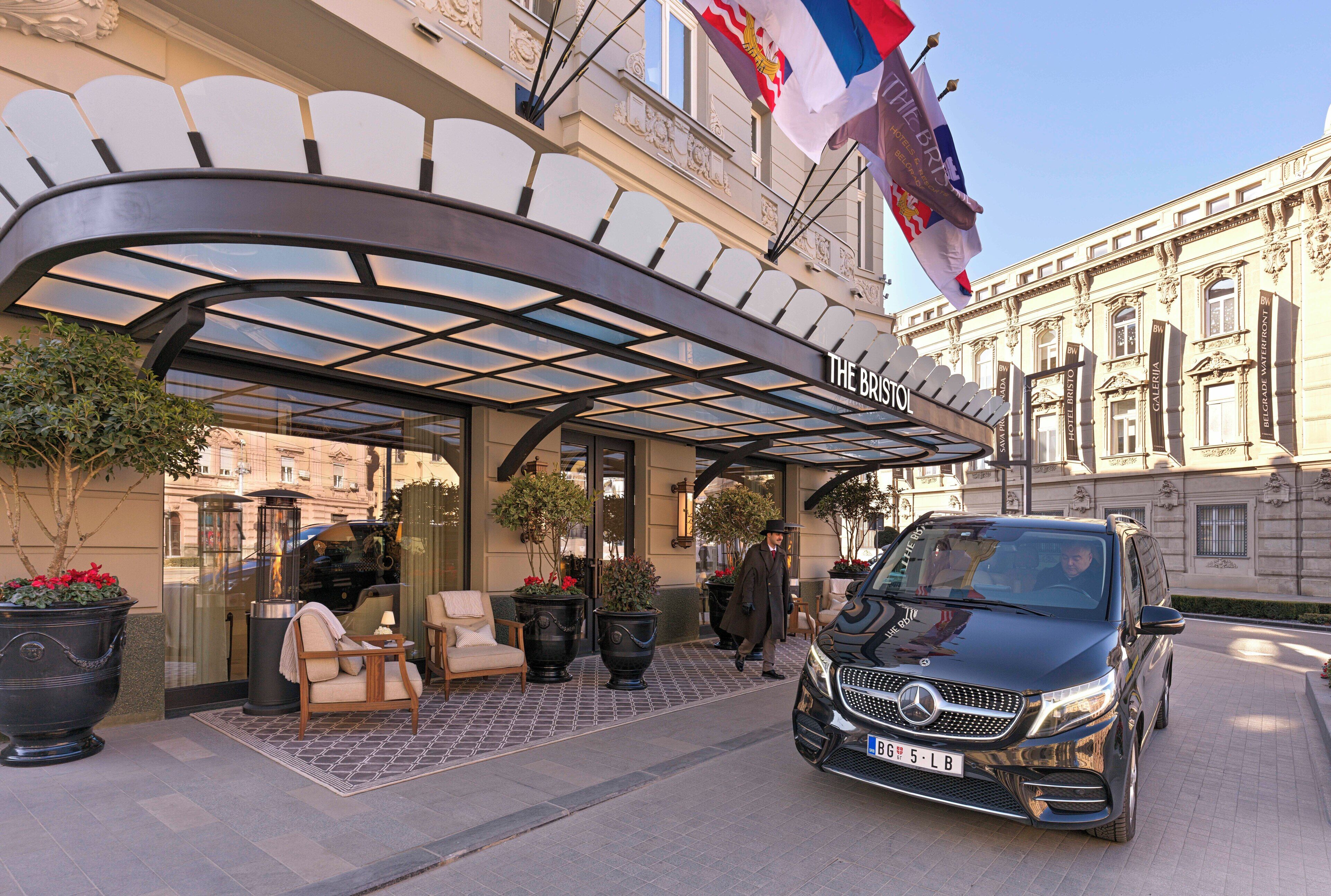 A hotel entrance with a covered outdoor seating area, a Mercedes-Benz SUV parked in front, and flags above.