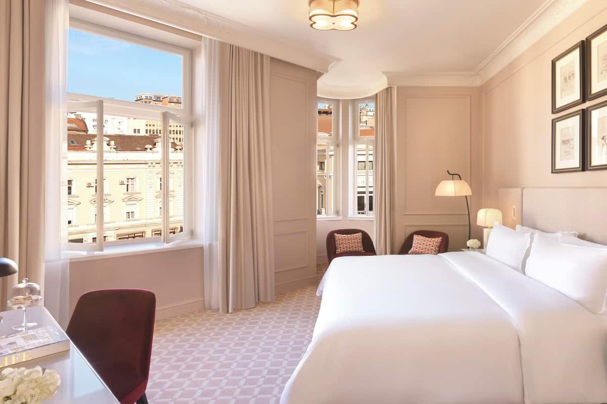 A hotel room with a large bed, a desk with a chair, a window with curtains, and framed pictures on the wall.