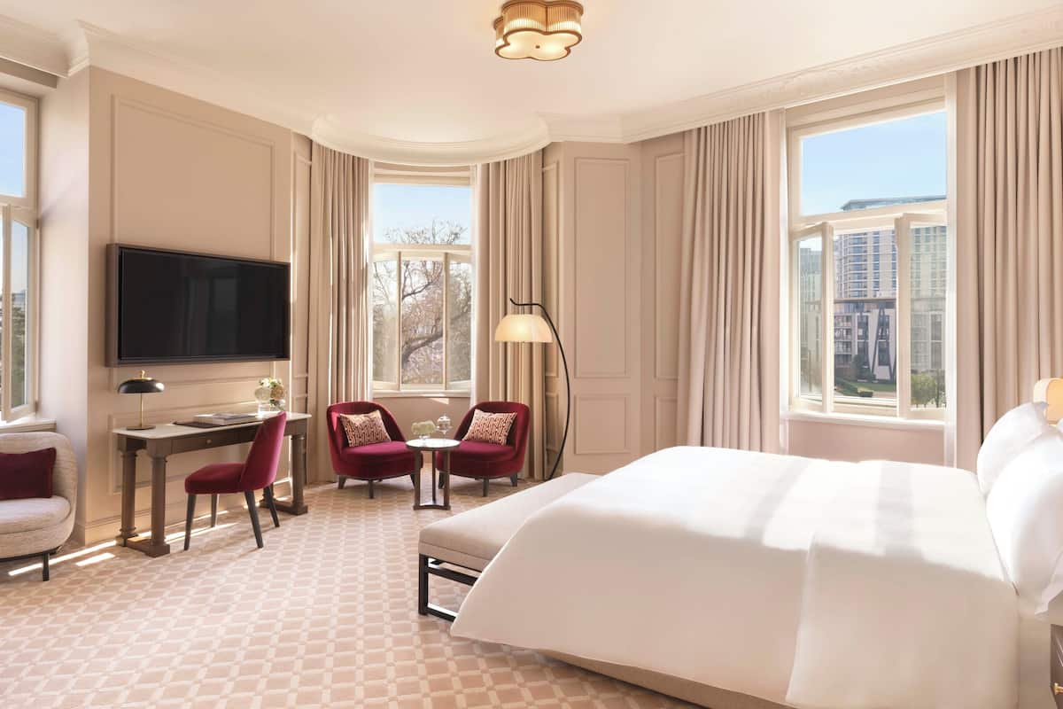 A hotel room with a large bed, a desk with a chair, two armchairs, a TV, and large windows with curtains.