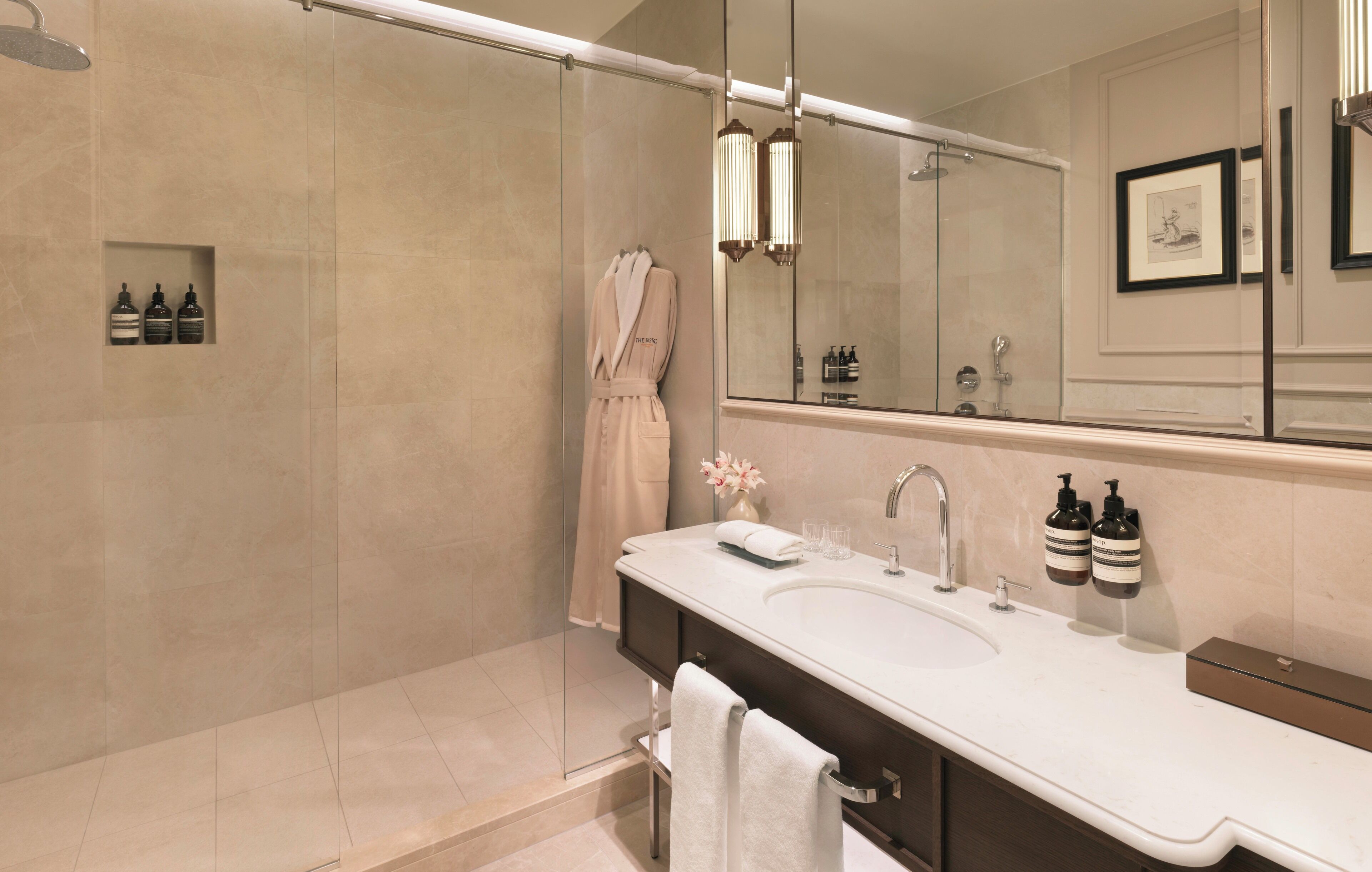 A modern bathroom with a glass-enclosed shower, a large mirror, a white sink with two bottles of shampoo, and a robe hanging on the wall.