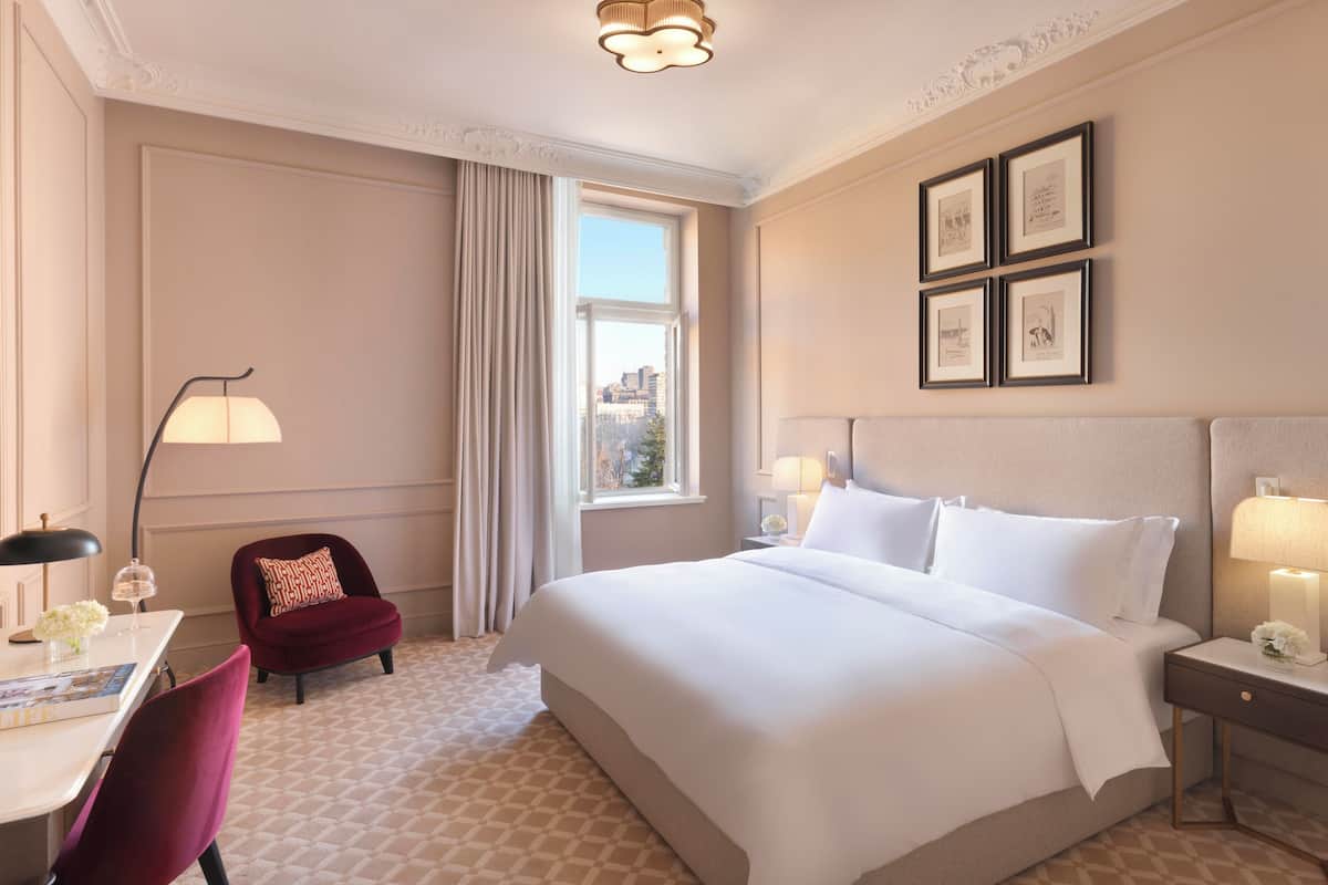 A hotel room with a large bed, a desk with a chair, a red chair, a nightstand with a lamp, and a window with curtains.