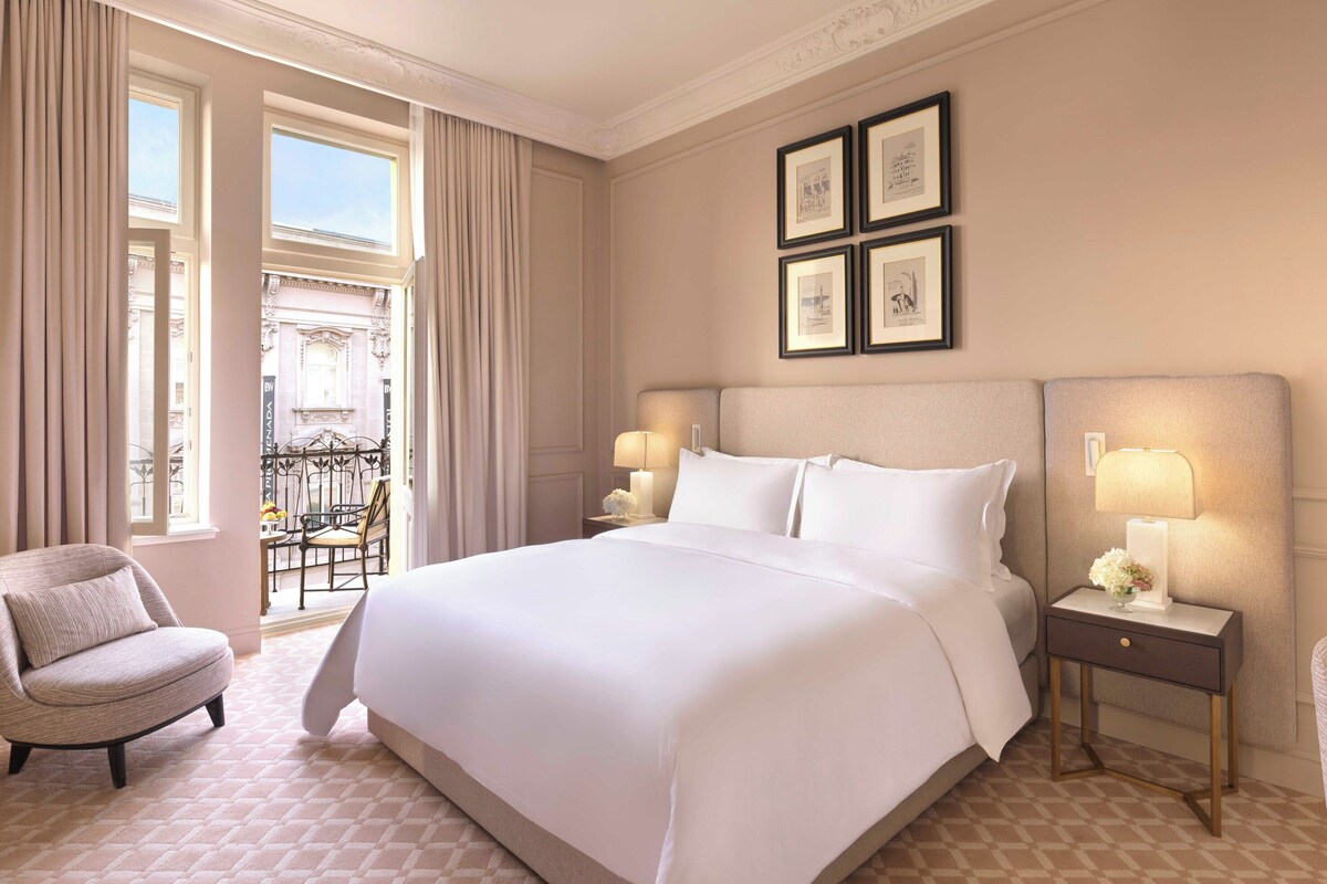 A hotel room with a large bed, two bedside tables with lamps, a chair, and a view of the outside.