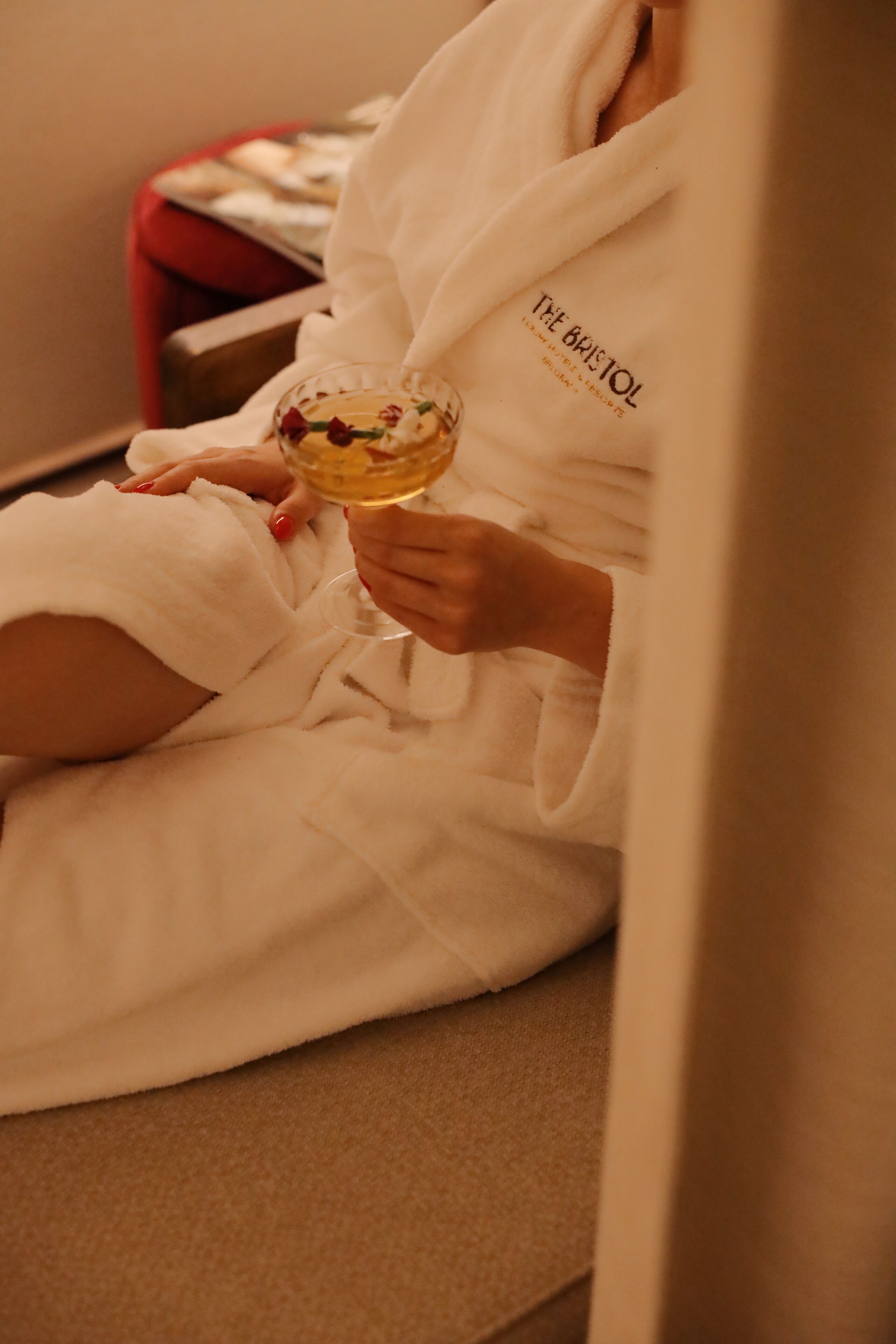 A person in a white bathrobe holding a glass of wine, seated on a beige couch with a red chair in the background.