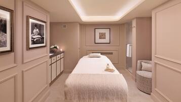 A spa room with a massage table, framed pictures, a small cabinet, and a patterned chair.