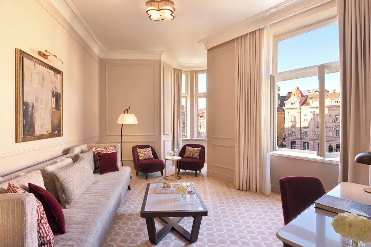 A hotel room with a sofa, armchairs, a coffee table, and a view of the city through large windows.