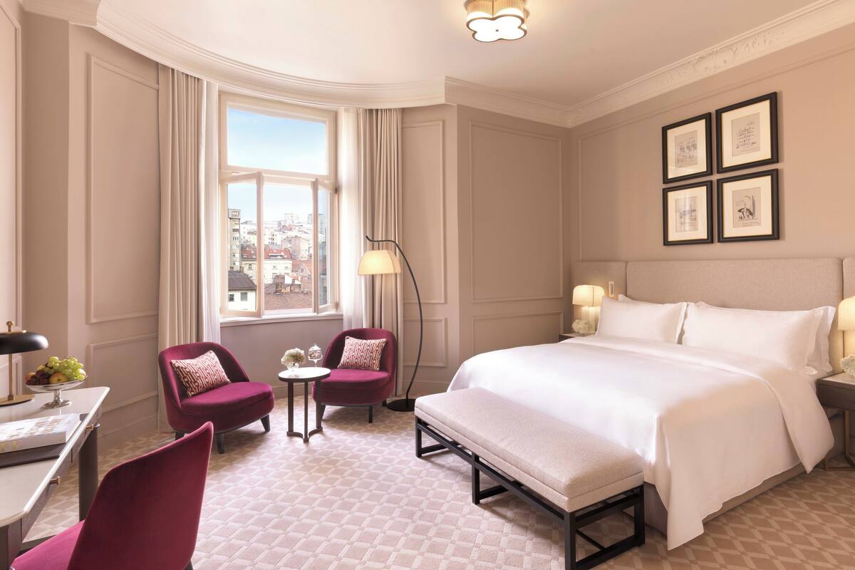 A hotel room with a large bed, two armchairs, a small table with a lamp, and a view of the city through a large window.