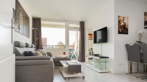 A modern living room with a grey sofa, a glass coffee table, a TV mounted on the wall, and a balcony with a view of buildings. - Vacation apartment "Peacock" in the recreation center Laboe (Laboe)