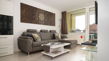 A modern living room with a grey sectional sofa, a glass coffee table, and a large window with curtains.