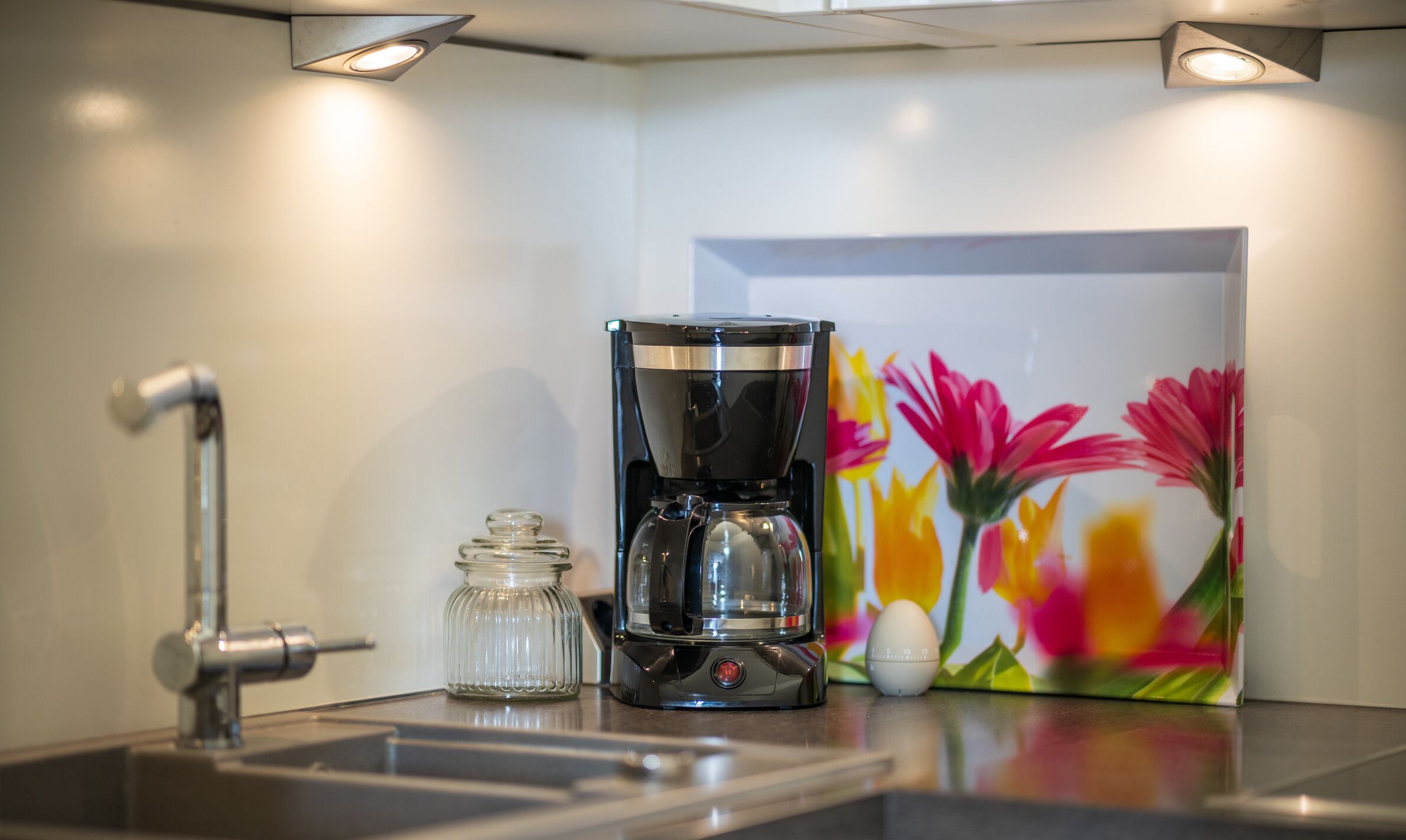 A kitchen with a coffee maker, a glass jar, and a colorful flower painting on the wall.