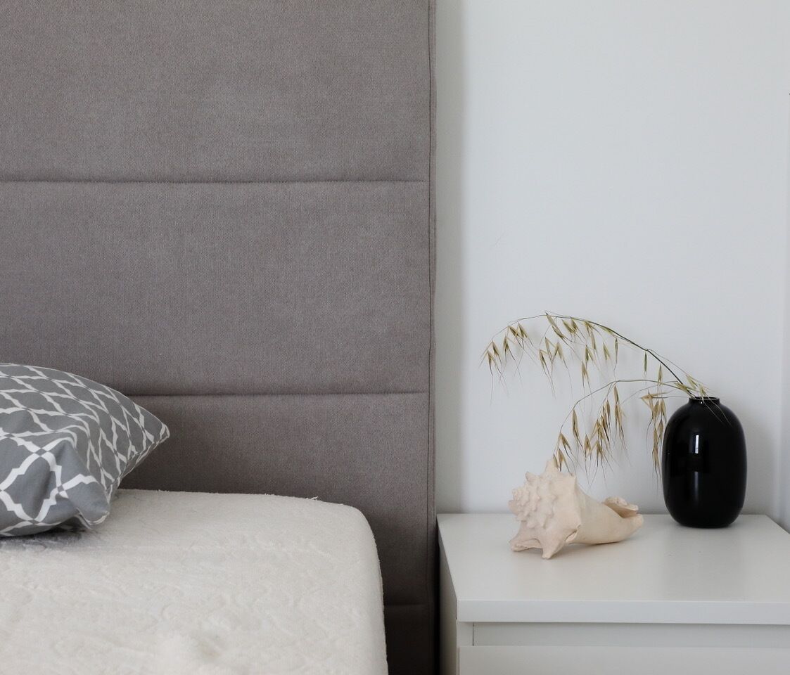 A neatly arranged bedroom with a grey upholstered headboard, a white bedspread, a patterned pillow, and a bedside table with a black vase and dried plants.