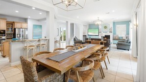Eine geräumige Küche mit einer zentralen Kochinsel, ein Essbereich mit Holztisch und Korbstühlen sowie ein Wohnzimmer mit Sofa und Fernseher.