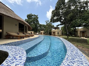 A swimming pool with blue mosaic tiles, lounge chairs, and a thatched-roof building.