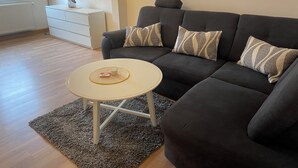 Una sala moderna con un sofá gris oscuro, dos cojines estampados, una mesita auxiliar blanca y una mesa de centro redonda con un vaso y una caja encima.