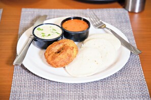 A plate with a white flatbread, a fried dough item, and two small bowls of soup on a woven placemat. - VILLA GRAND HOTEL NEAR BENGALURU AIRPORT (Bengaluru)
