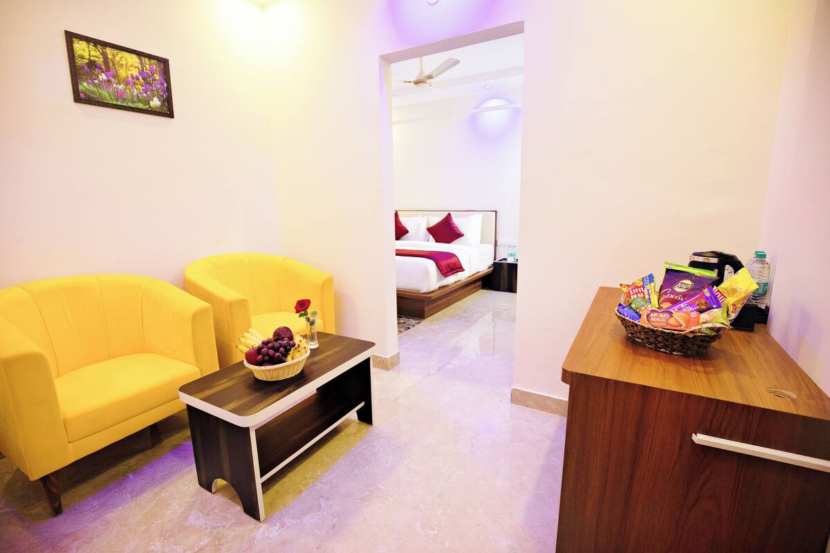 A hotel room with a yellow sofa, a small table with a fruit bowl, a wooden desk with snacks, a bed with red pillows, and a ceiling fan.