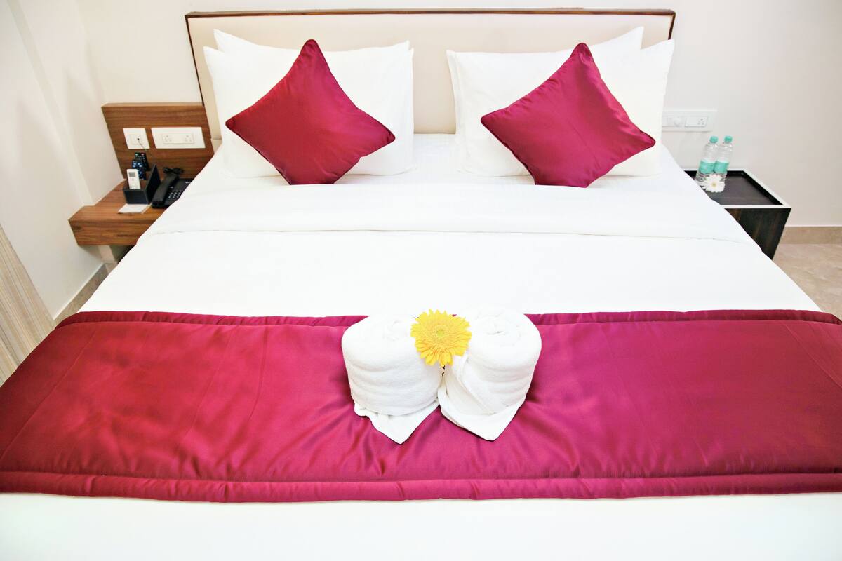 A neatly made bed with white and red bedding, a wooden nightstand with a phone and remote, and a small table with water bottles.