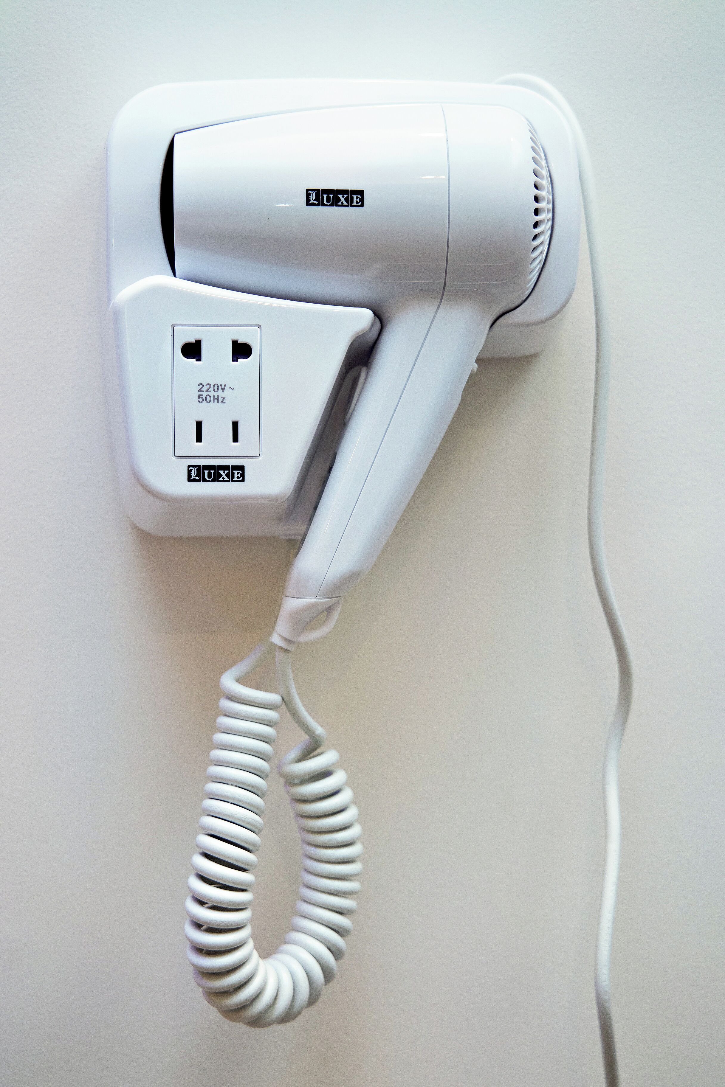 A white hair dryer with a coiled cord, mounted on a wall with a power outlet.