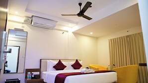 Een hotelkamer met een bed, een plafondventilator, een airconditioner, een spiegel, een nachtkastje met een telefoon en een fles, en een gele fauteuil.
