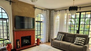 Una sala de estar con chimenea, un sofá, un televisor y ventanas grandes con cortinas.