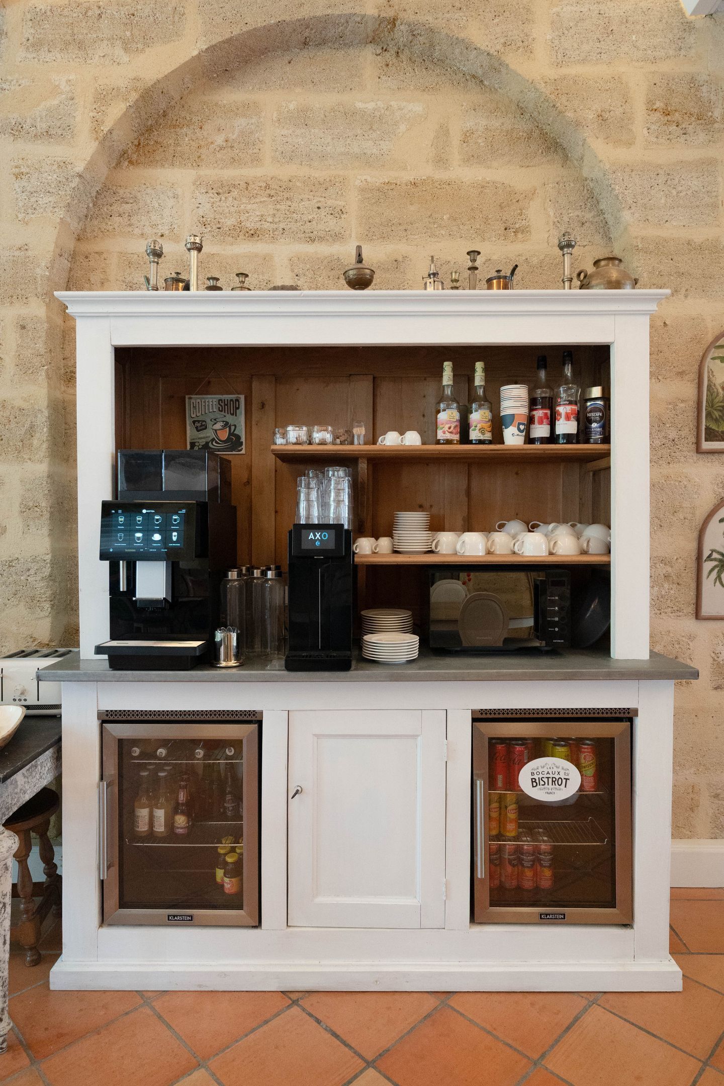 Mostrador de cafetería con máquina de café, estantes con tazas y botellas, y un refrigerador con bebidas.