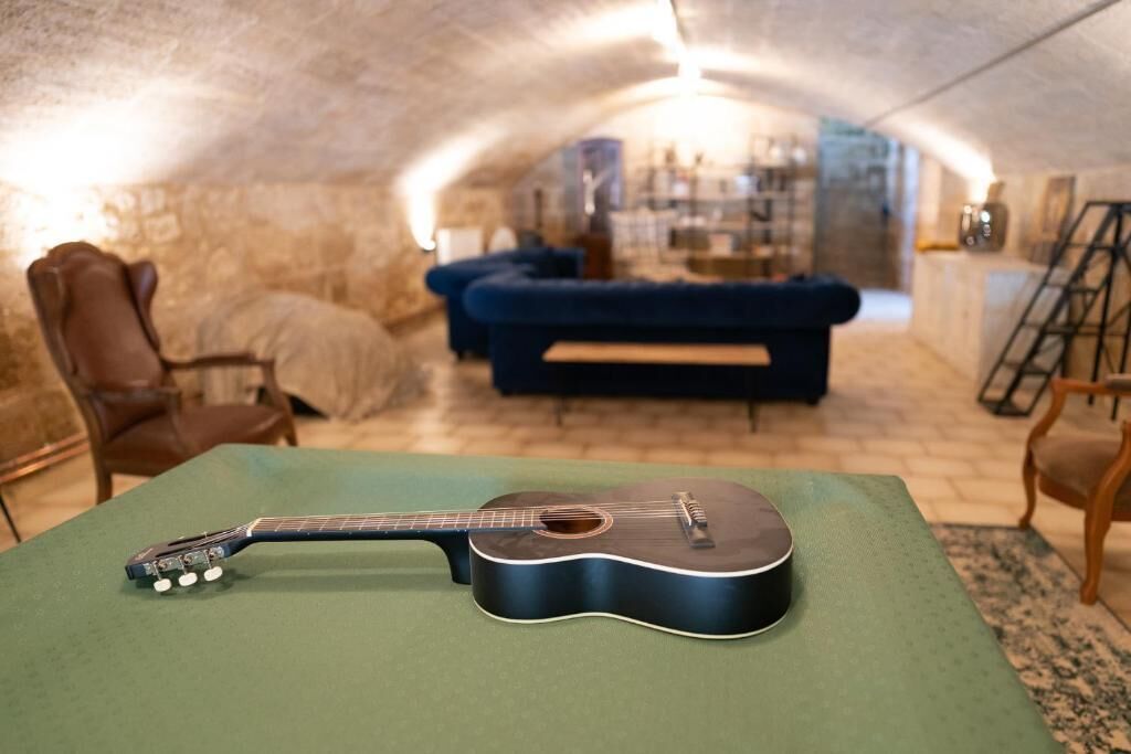 Une guitare posée sur une table verte, dans une pièce chaleureuse aux murs de pierre, avec un canapé bleu et des chaises en bois.