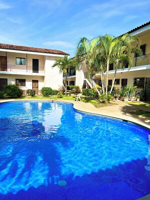 A swimming pool surrounded by a courtyard with a building featuring balconies and a tiled roof.