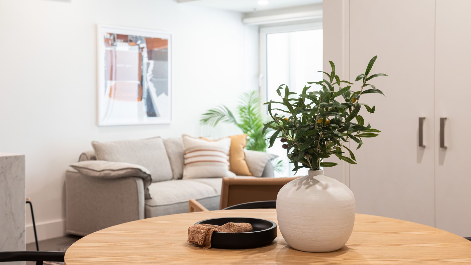 Ein modernes Wohnzimmer mit einem runden Holztisch, einer weißen Vase mit Grünpflanzen und einer schwarzen Ablagefläche.