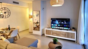 A modern living room with a dining area, a TV displaying a mobile app, and a large clock on the wall. - Luxury 2-Bedroom Apt with Jacuzzi, Patio & Parking 1km from Beach (Torremolinos)