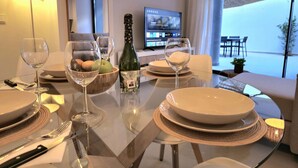 A dining table set with plates, glasses, and a bottle, with a TV in the background and a view of the outdoors. - Luxury 2-Bedroom Apt with Jacuzzi, Patio & Parking 1km from Beach (Torremolinos)