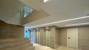 Ein modernes Interieur mit einer Treppe, die zu einer oberen Ebene führt, einem Gang mit drei Aufzügen und einer Tür am Ende.