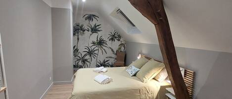 Un dormitorio con techo inclinado, una cama con colcha color beige, dos almohadas y una mesita de noche.