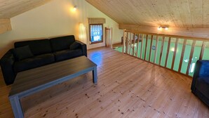 Een kamer met houten vloer, een bank en een tafel, een raam met gordijnen en uitzicht op een groen gebied buiten.