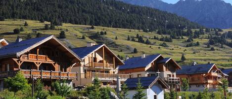 A row of wooden chalets with sloped roofs, set against a backdrop of a mountainous landscape.