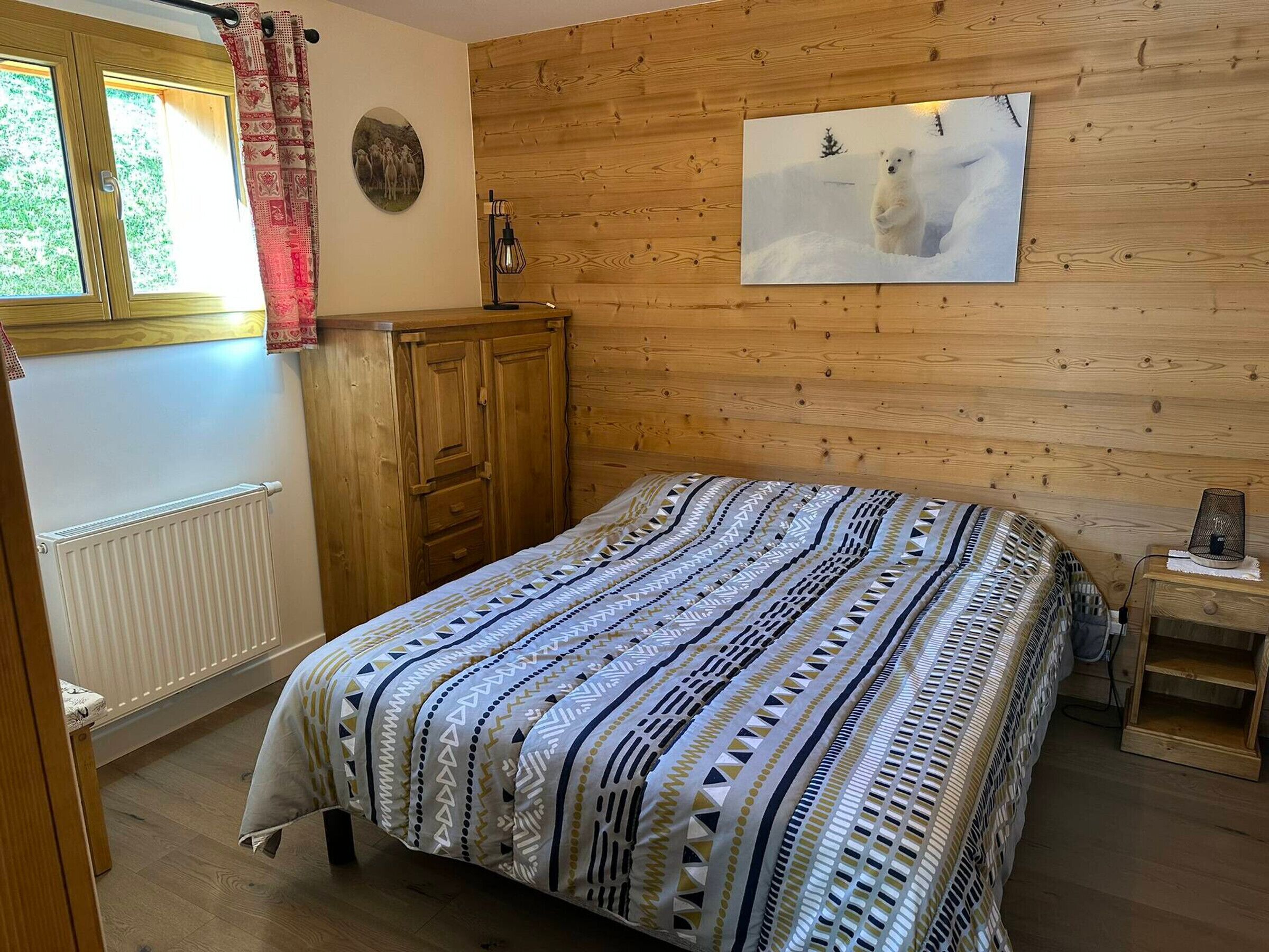 Une chambre à coucher avec une tête de lit en bois, un lit recouvert d’une couvre-lit à motifs, une armoire en bois, un radiateur et une fenêtre avec des rideaux.