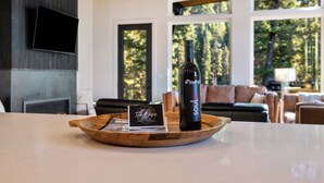 A wooden tray with a bottle of wine and a business card on a countertop in a modern living room with a fireplace, large windows, and a television mounted on the wall. - The Basin is a modern, luxury cabin nestled in the pines at the base of Discovery Ski Hill. Enjoy panoramic views of the surrounding forest and the relaxing sounds of a nearby creek while relaxing by a smokeless fire in the Solo stove on the deck. (Philipsburg)