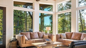 A spacious living room with two beige sofas, a coffee table, and a view of a forest through large windows. - The Basin is a modern, luxury cabin nestled in the pines at the base of Discovery Ski Hill. Enjoy panoramic views of the surrounding forest and the relaxing sounds of a nearby creek while relaxing by a smokeless fire in the Solo stove on the deck. (Philipsburg)