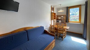 A cozy room with a blue sofa, wooden dining table, and chairs, a TV mounted on the wall, and a window with a view of buildings.