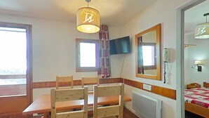 A room with a wooden dining table and chairs, a ceiling lamp, a TV mounted on the wall, a bed with a quilt, and a radiator. - Apartment 5 pers, parking, Plagne Soleil (Plagne Soleil)