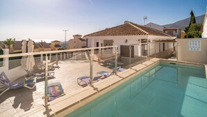 Un espace piscine aménagé sur le toit, avec des chaises longues, une rambarde en verre et une vue sur les bâtiments et les montagnes environnants.